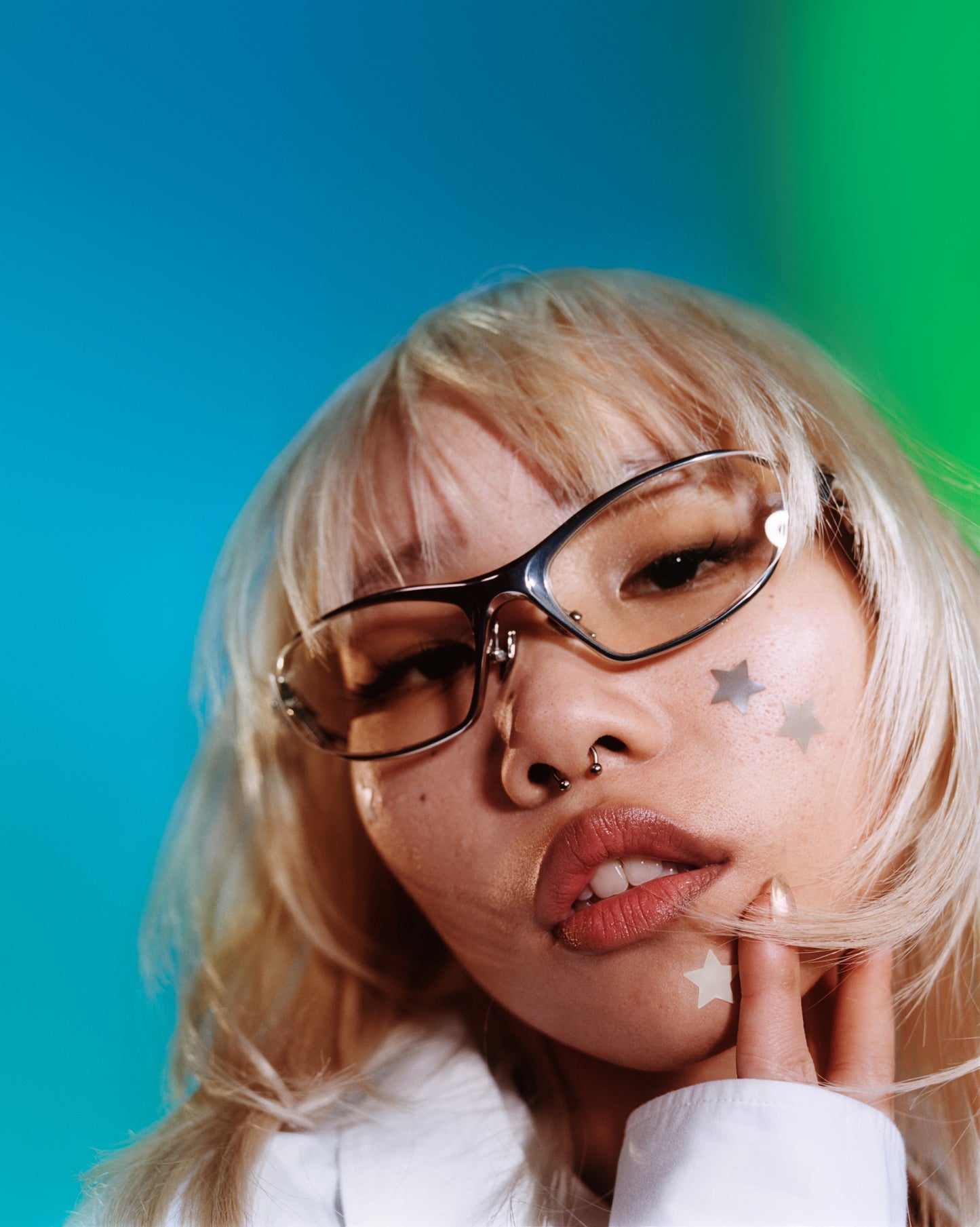 Young blonde model in chrome glasses wears two neutral hydrocolloid Earth stars on her cheek and one Earth star on her chin with her hand on chin.