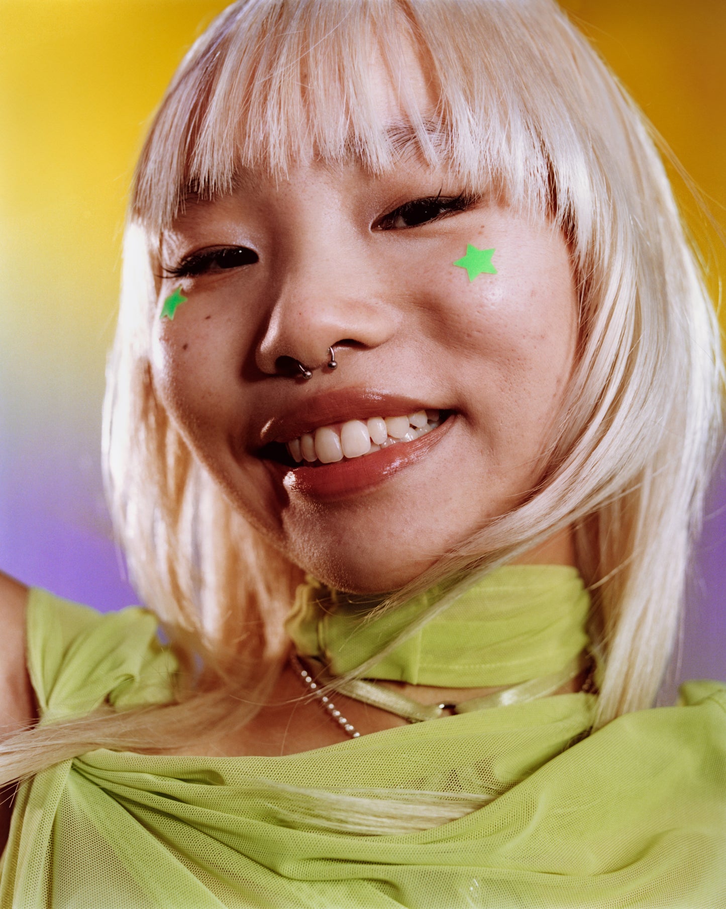 A young smiling person with blonde hair wears two green Starface Hydro-Star® + Tea Tree star-shaped patches on their cheeks.