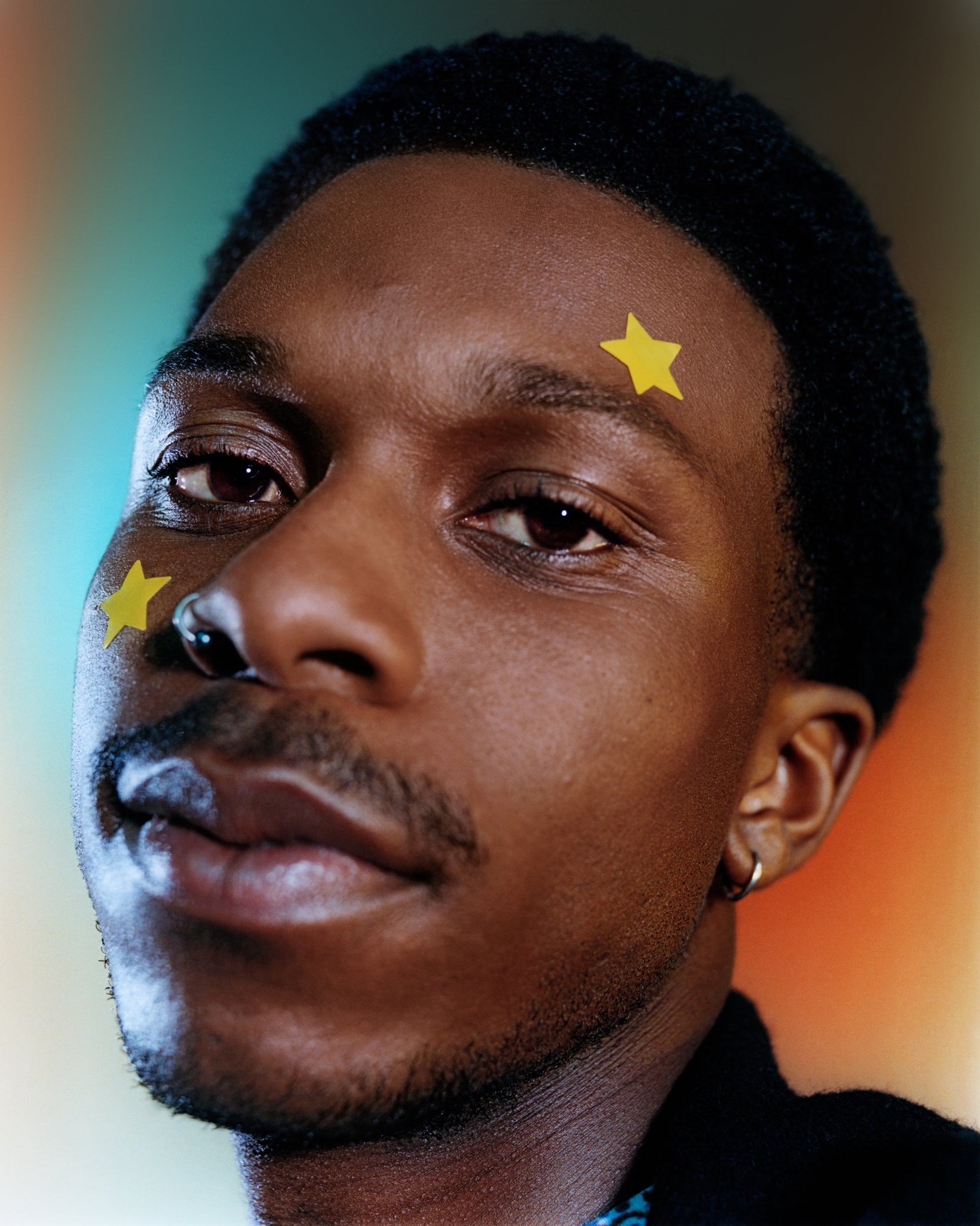 A young person with a mustache wears a yellow XL Big Star hydrocolloid  pimple patch above their eyebrow and on their cheek.