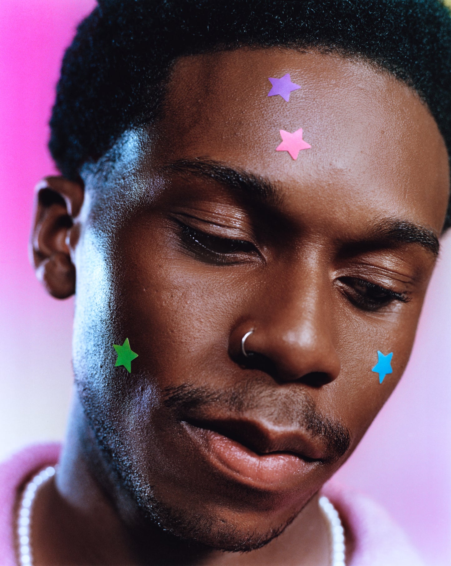 Young person with a nose ring wears blue, green, pink, and purple star-shaped Party Pack hydrocolloid pimple patches on their face.