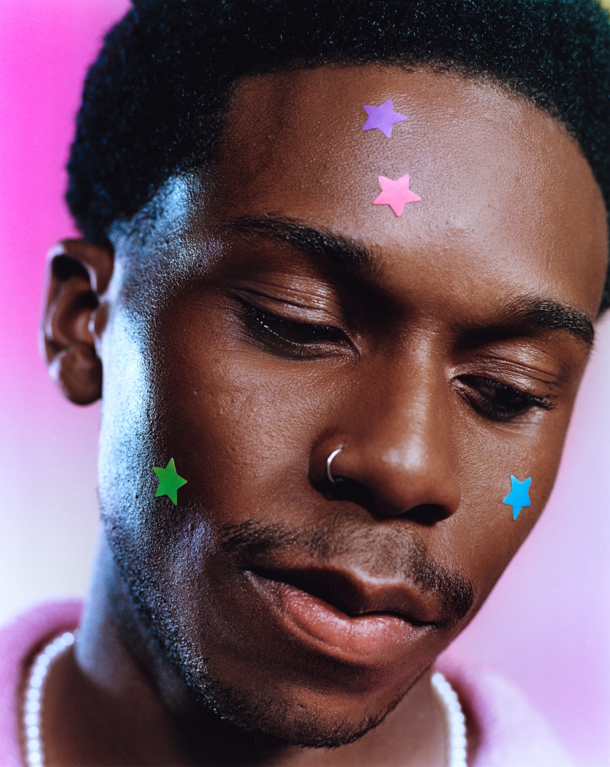 Young person with a nose ring wears blue, green, pink, and purple star-shaped hydrocolloid pimple patches on their face. 