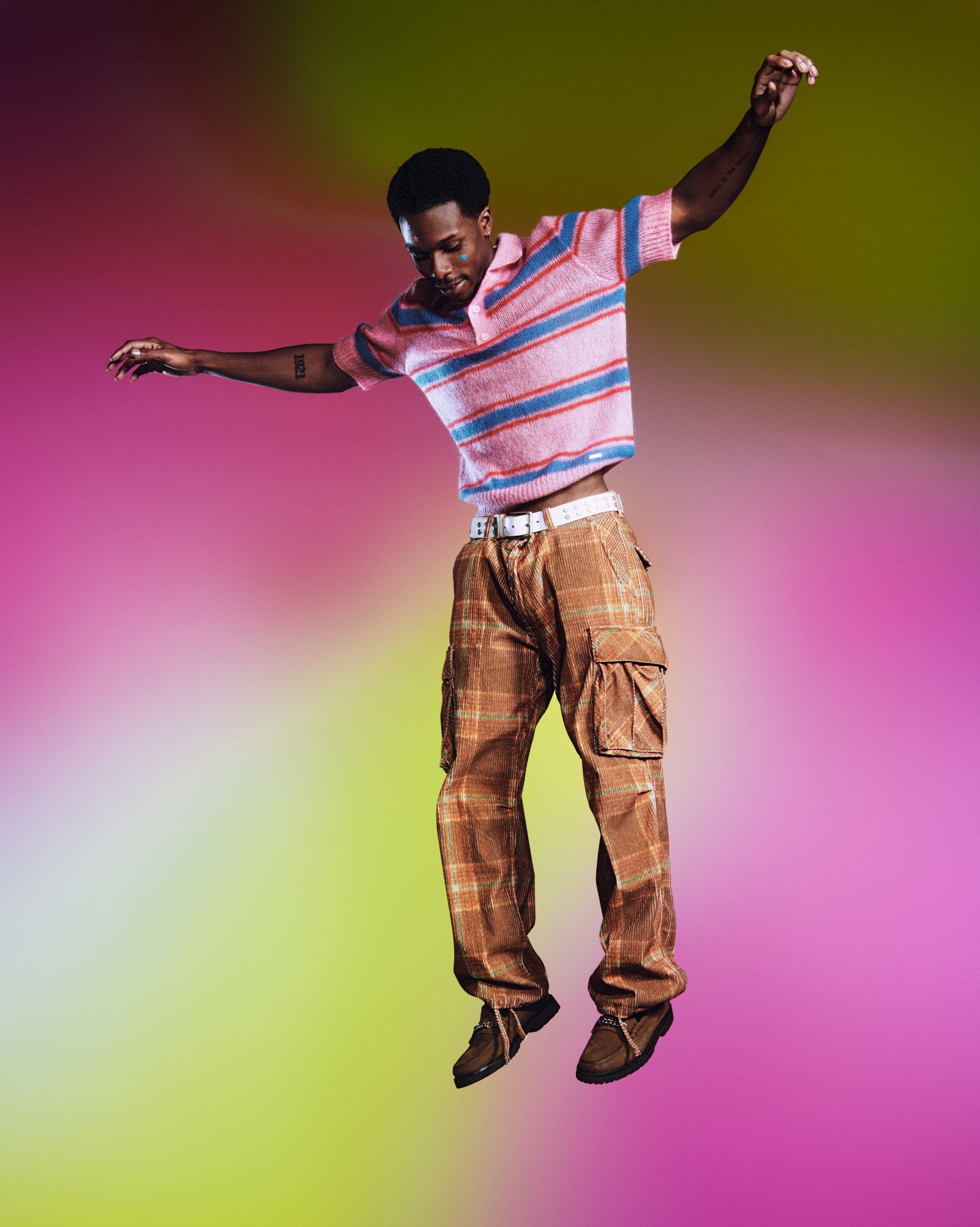 Model in a pink shirt, orange pants, and a white belt floats in the air while wearing colorful Party Pack hydrocolloid pimple patches. 