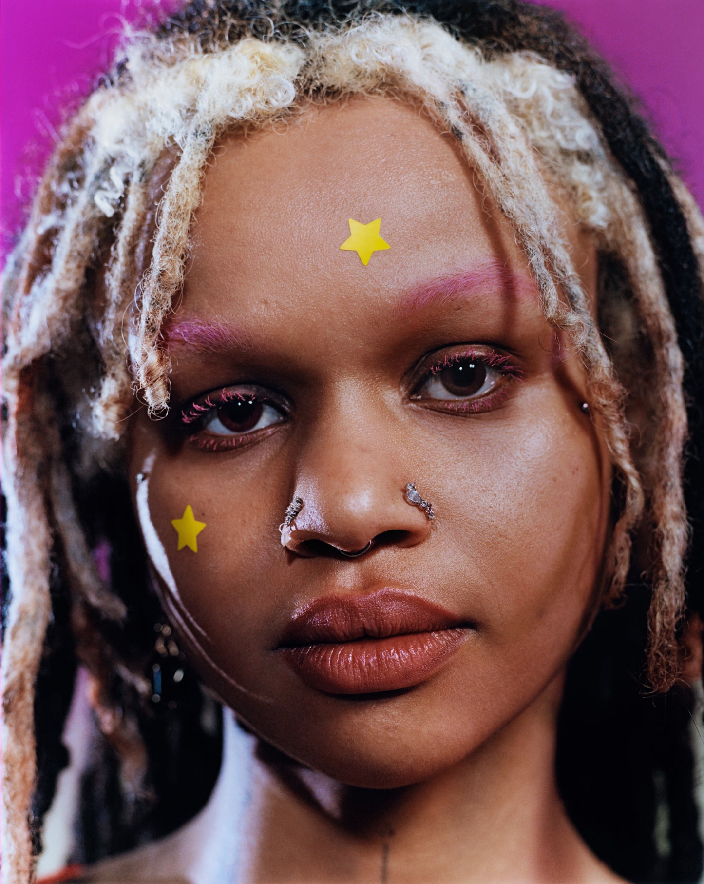 A young person with white and black hair, pink eyebrows, and two nose rings wears yellow Hydro-Star® patches on their cheek and forehead.